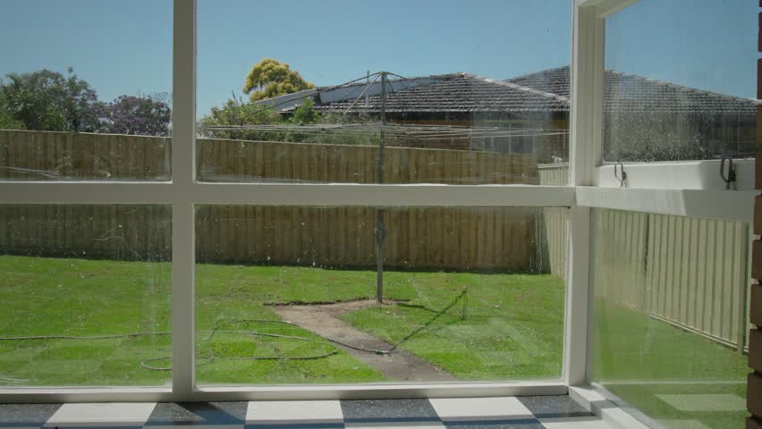 View from inside a residential home looking out through a slightly dirty window
onto a suburban backyard, where a sprinkler waters a green lawn. With a classic Australian hill hoist clothesline.