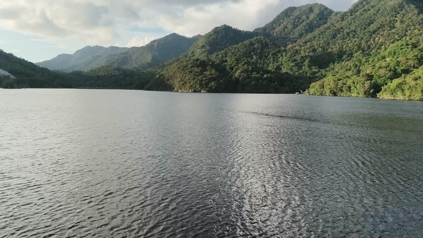 A wide, tranquil mountain lake is surrounded by lush, green, tree-covered hills under a dramatic cloudy sky, showcasing a serene natural landscape.