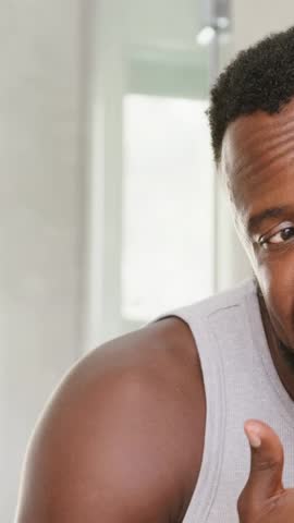 Vertical video: Leaning to mirror, man rubbing cheek, stroking beard in bathroom, wearing tank top. Grooming, skincare, closeup, vanity, masculine, adult, vertical video