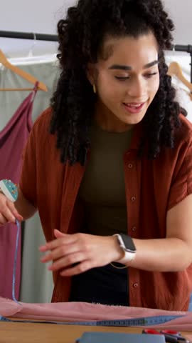 Vertical video: Reading tape, woman in rust top measuring fabric for sewing and cutting in studio. Seamstress, garment, textile, tailoring, scissors, pincushion, vertical video