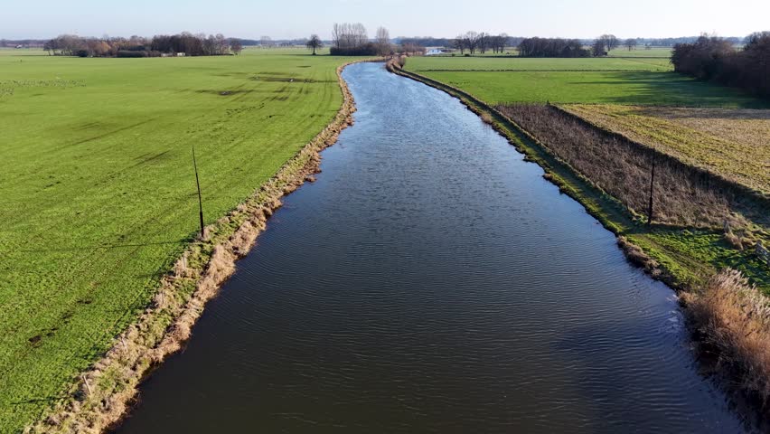 An aerial view of a wide river flowing through vibrant green fields. A large flock of birds flies across the water and meadows under a clear sky, capturing a serene rural European landscape.