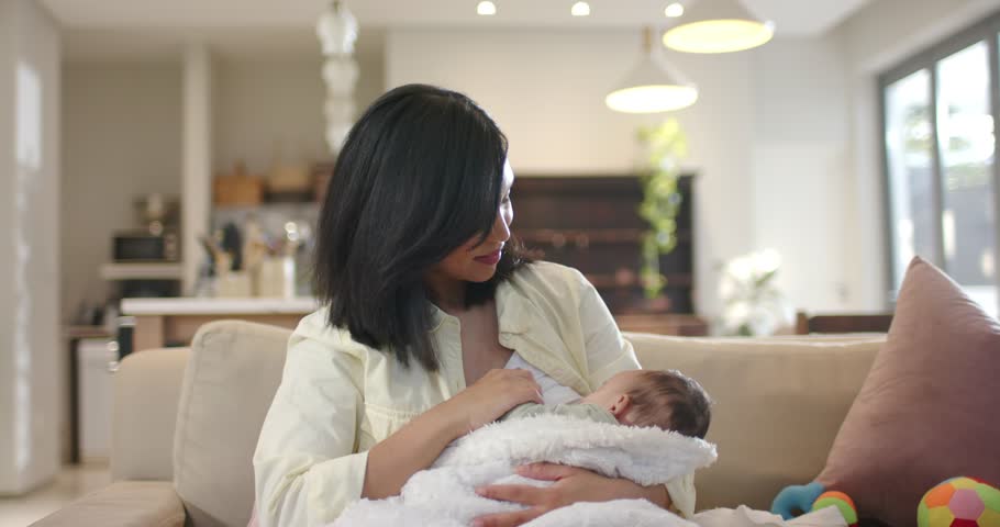 Mother lifting baby to chest for feeding while stroking head, adjusting grip on sofa near window. Parenting, nurturing, intimacy, comfort, minimalist, cozy, family