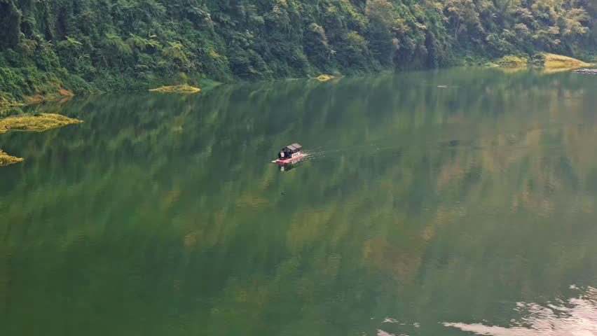 Person paddles small boat across calm green lake. Forested hills surround still water surface during daytime.