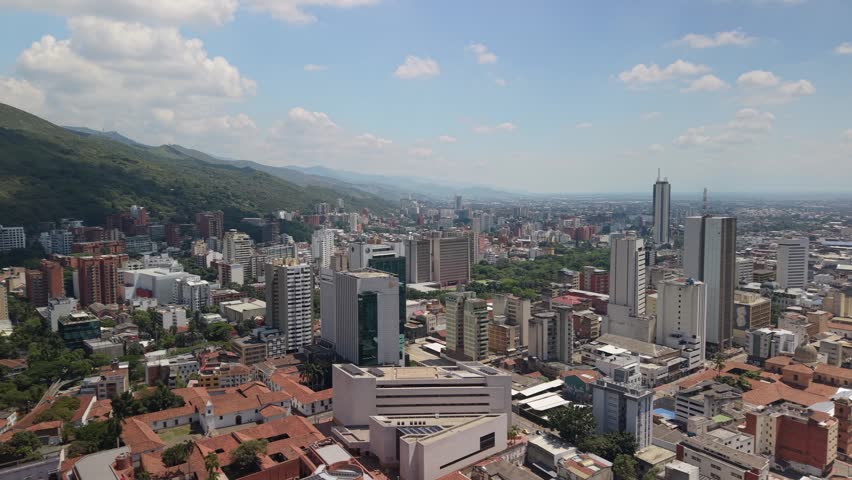 Drone orbit sequence revealing the lush Farallones mountains and downtown skyline of Cali. View includes colonial rooftops, modern office buildings, and the iconic Torre de Cali tower.