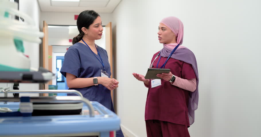 Two adult female nurses in scrubs responding to tablet update, discussing patient care in corridor. Teamwork, hospital, clinical, gesturing, device, stethoscope, badge