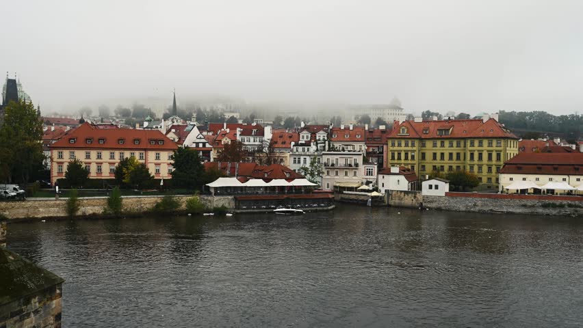 Historic Prague buildings line Vltava River, morning view from Charles Bridge. Misty conditions shroud the city.