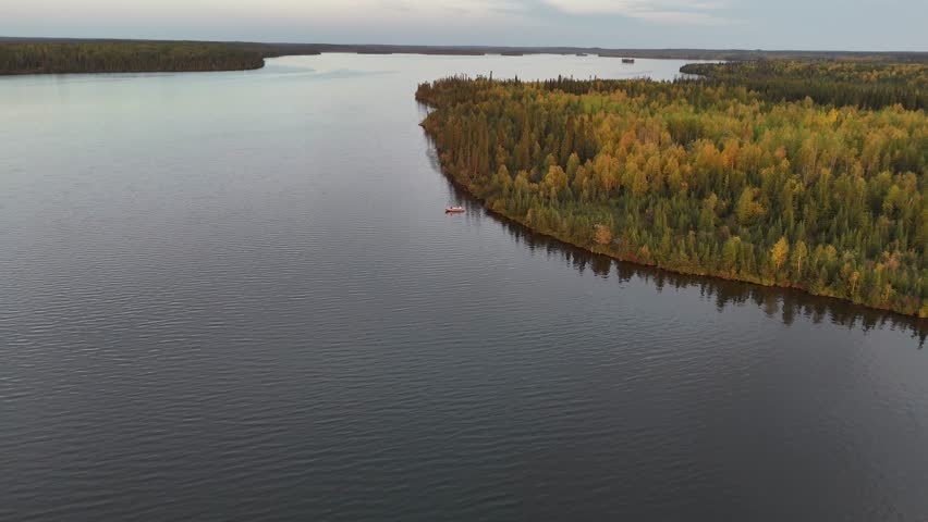 Aerial drone descends and moves toward a small fishing boat on a lake in northern Manitoba, Canada. Boreal forest with fall colors surrounds calm water in natural light.