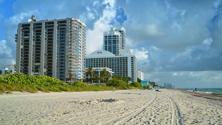 Sandy beach with tire tracks and high-rise buildings in Miami. Wide shot of North Miami Beach coastline with vehicle tracks in sand and residential skyscrapers under cloudy sky.