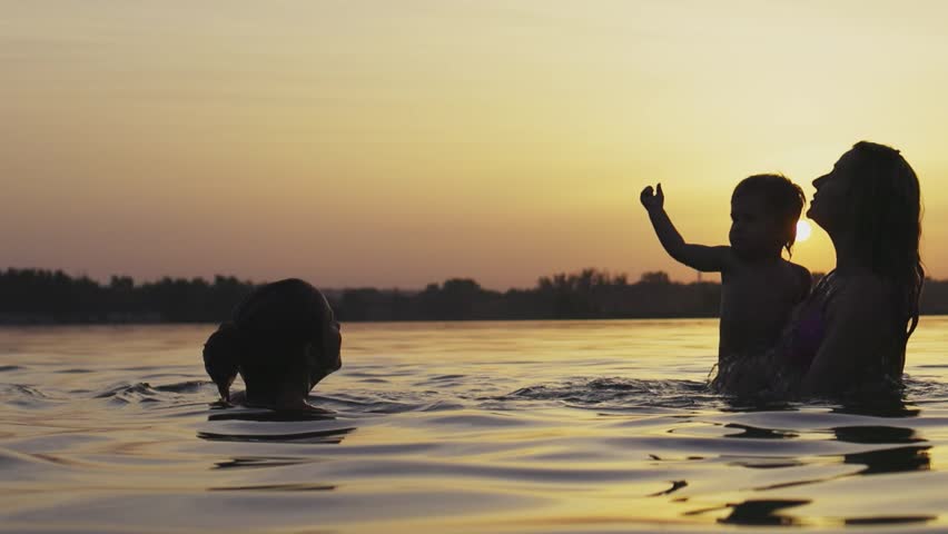 Mom with the baby she holds in her arms playing with her daughter and splashing in the lake against background of sunset