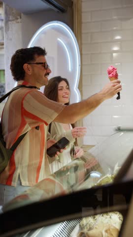 Young man and woman on a date choosing different flavors from an ice cream parlor display case. Happy tourists enjoying a cold summer dessert