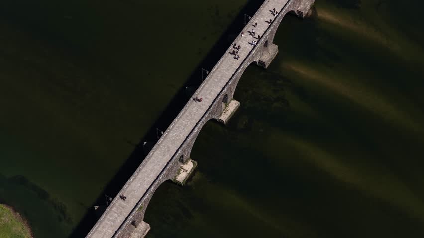 Aerial view of an ancient stone bridge with people crossing it, casting a long shadow over the water, Ponte de Lima, Portugal.