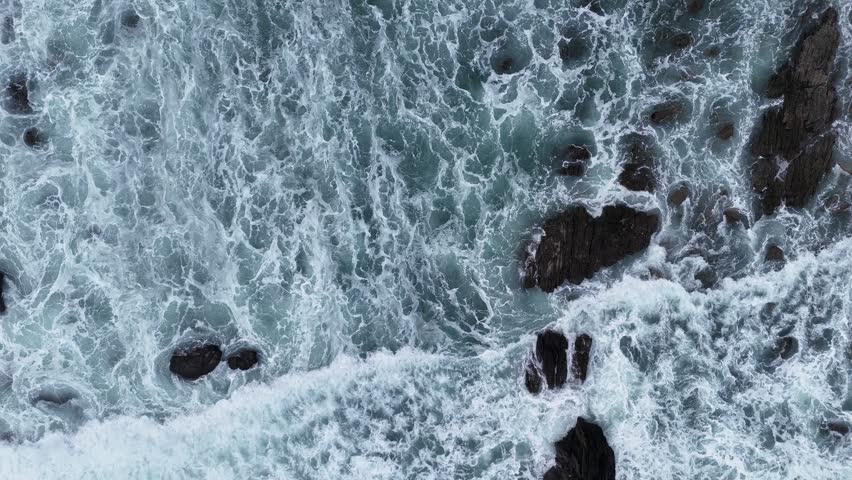 Vertical aerial shot of foamy waves washing over dark rocks along a rugged shoreline.