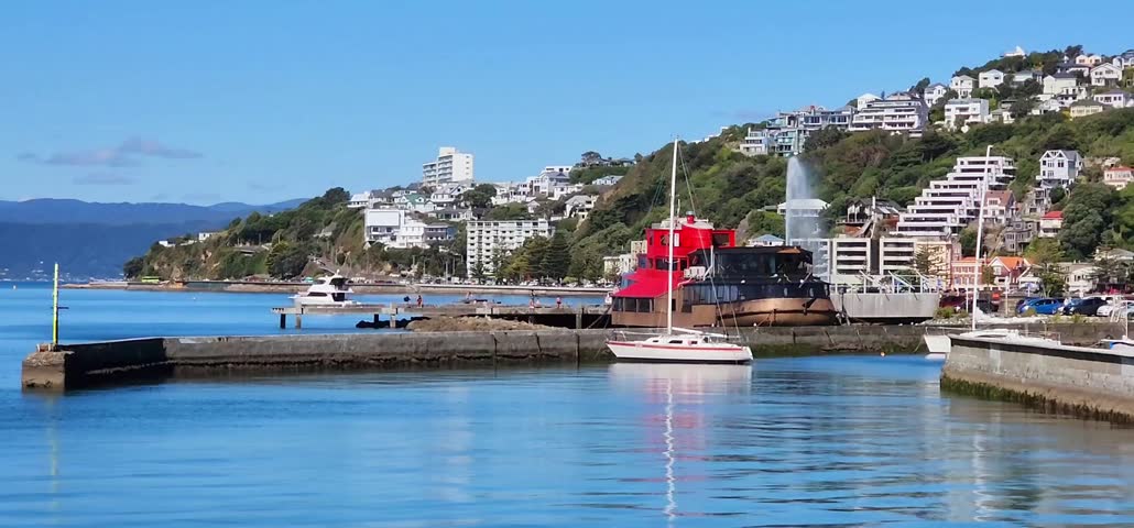 Wellington, New Zealand - 22 January 2025: Harbourfront Boats and Hillside Cityscape with Fountain on a Clear Day. A waterfront scene in Wellington, New Zealand, featuring boats docked near concrete piers and a distinctive red vessel with a water fountain spraying behind it. White hillside buildings rise above the harbour, framed by green slopes and a clear blue sky. The calm water and colourful coastal architecture highlight Wellington’s blend of maritime activity and scenic urban development. This video is suitable for travel journalism, harbour features and New Zealand‑focused contents.
