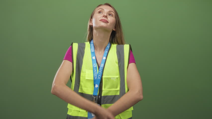 Woman volunteer in reflective vest with blue lanyard touches chin in a thinking pose at studio; thoughtful service.