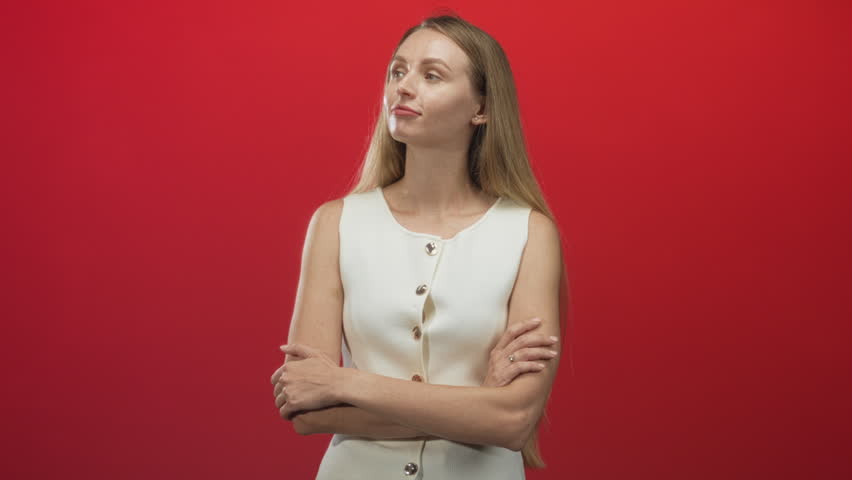 Woman with arms crossed and a slight smile wearing a white sleeveless buttoned top against a red studio backdrop; confidence.