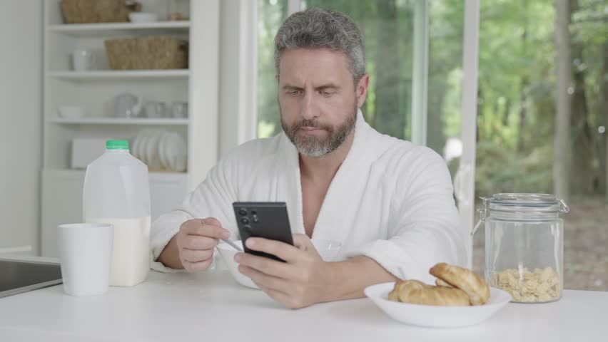 Man eat breakfast in the kitchen. Guy eating cereal with milk. Hispanic man enjoy healthy meal at kitchen table. Healthy nutrition mature man. Aged man with healthy products at kitchen with phone