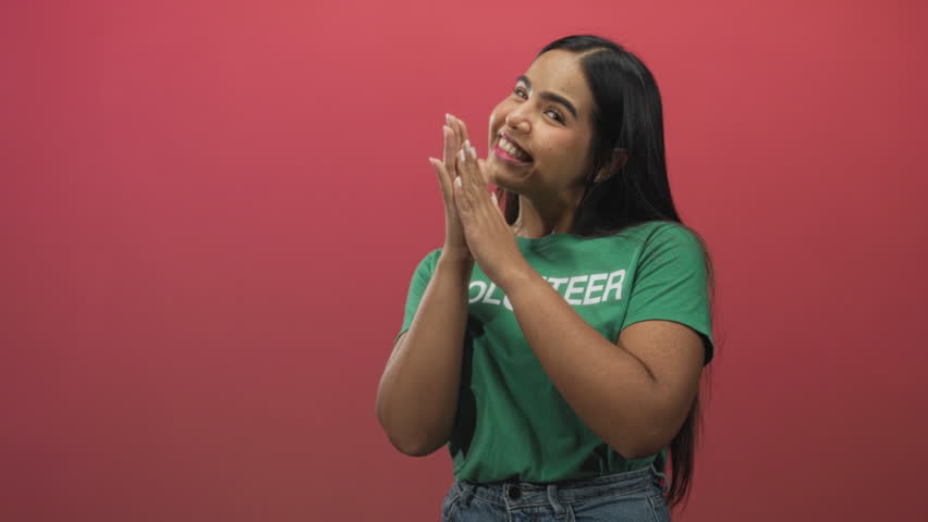 Young volunteer woman wearing green tshirt and denim jeans clasping hands near face and smiling in studio; gratitude warmth.