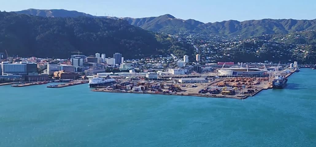 Harbourfront City Skyline with Port Facilities and Forested Hills. A panoramic view of Wellington, New Zealand, showing the city’s waterfront skyline, port facilities and dockside infrastructure along the harbour. High‑rise buildings cluster near the shoreline while forested hills rise behind the urban centre. The vivid blue water of Wellington Harbour contrasts with the industrial piers and the surrounding natural landscape. Recommended for travel journalism, harbour documentation and New Zealand‑focused contents.