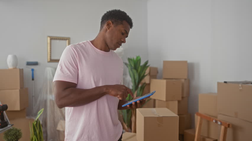Man standing tapping tablet with finger amid moving boxes, ladder, drill and houseplant in building; focused planning.
