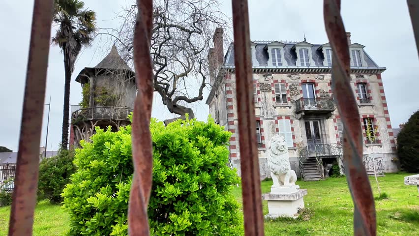 Panning shot of a grand historic French mansion with stone lion statues and a rustic wooden gazebo in the garden.