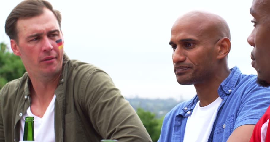 Diverse men at picnic table, painted man reacting, talking while Indian man eating burger. Outdoor, casual, friends, garden, dining, sandwich, salad