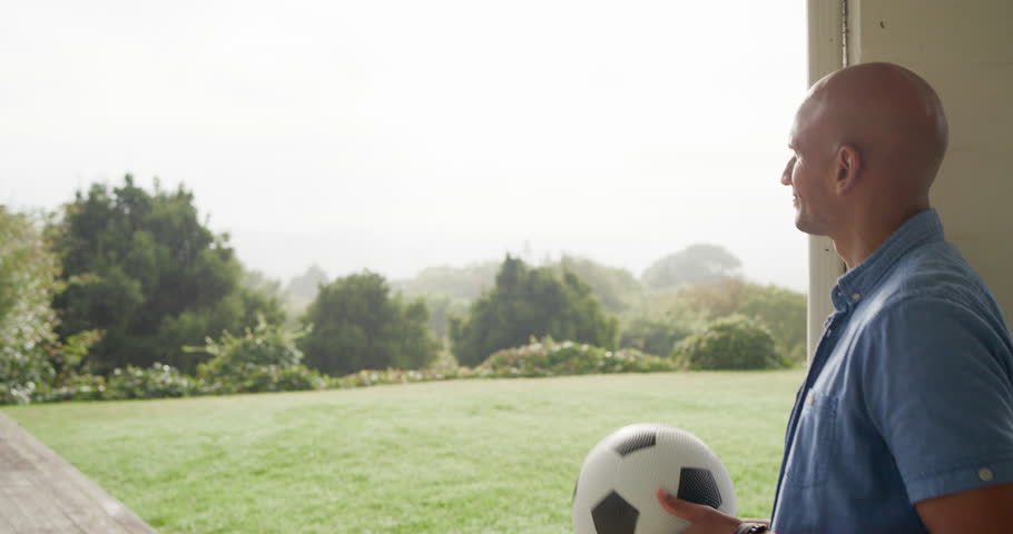 African American man standing on porch holding soccer ball, smiling at memory, copy space. Outdoor, doorway, yard, hills, candid, natural, serene