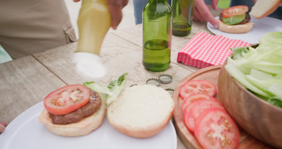 Vertical video: Diverse male friends hungry, making burgers at picnic table, passing mustard. Outdoors, barbecue, grilling, casual, gathering, greenspace, condiments