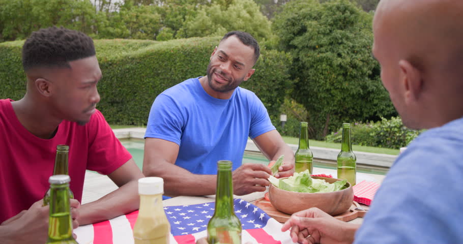 African American men sitting at pool barbecue flag cloth, man in blue tee holding lettuce, talking. Backyard, outdoor, patio, gathering, friends, grill, beverages