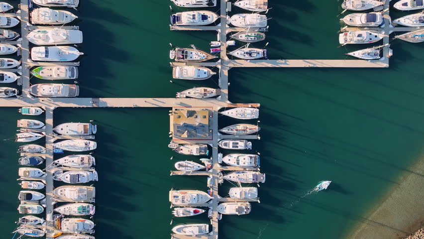 Drone view of harbor docks. Boats anchored in blue water. Marina layout from above. Clean geometric marina pattern. Yachts parked at the piers. Drone flight over marina. Calm harbor with boats.