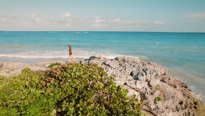 Woman walking along rocky shore by turquoise ocean waters in Tulum, Mexico