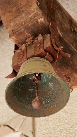 Close-up of aged rusty bronze bell hanging on old wooden structure in traditional Italian village. Historic rural detail with weathered patina and texture.