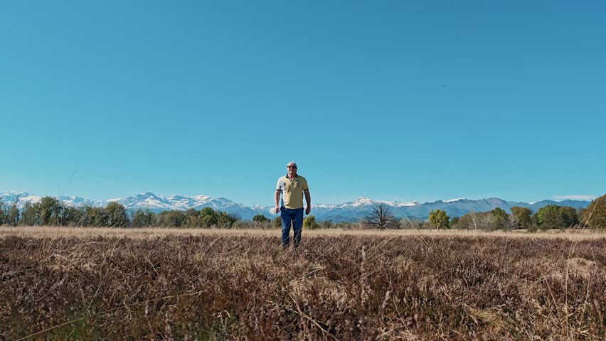 Happy man jumping with arms raised on open field with majestic snowy Alps in background. Carefree vacation mood under clear blue sky in northern Italy.