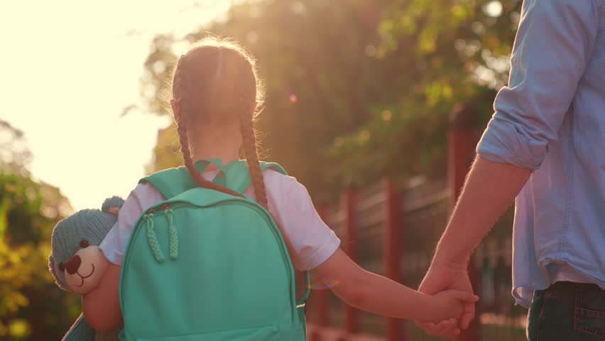 Dad parent, child girl, walking together outdoors. Father little schoolgirl daughter walking to school with backpack, child holding teddy bear. Children