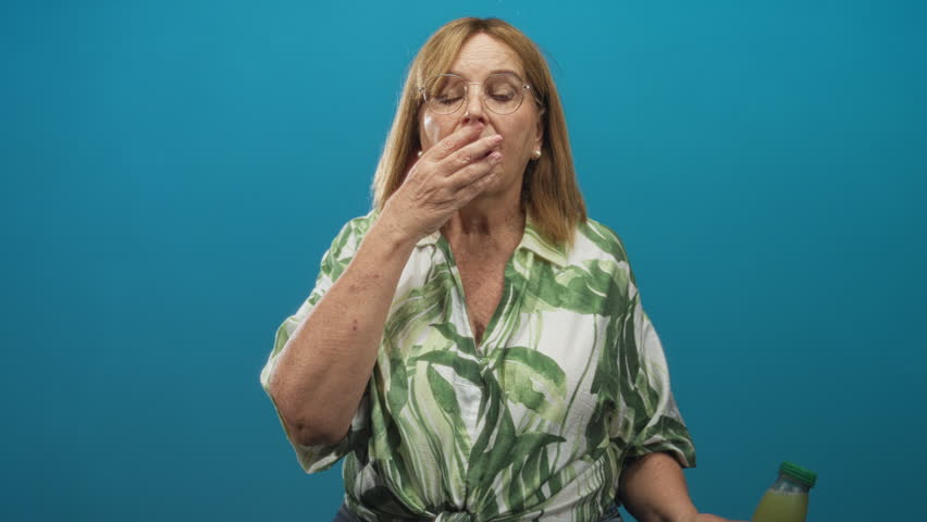 Woman holding green juice bottle with hand covering mouth in studio wearing a leaf print shirt and glasses; surprise wellness curiosity.