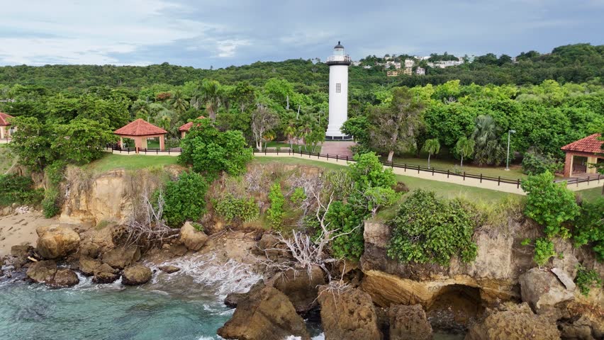 Aerial drone view of the Rincon lighthouse standing on a coastal cliff surrounded by tropical vegetation and overlooking the ocean in Puerto Rico. Scenic landscape featuring rocky shoreline and greenery.