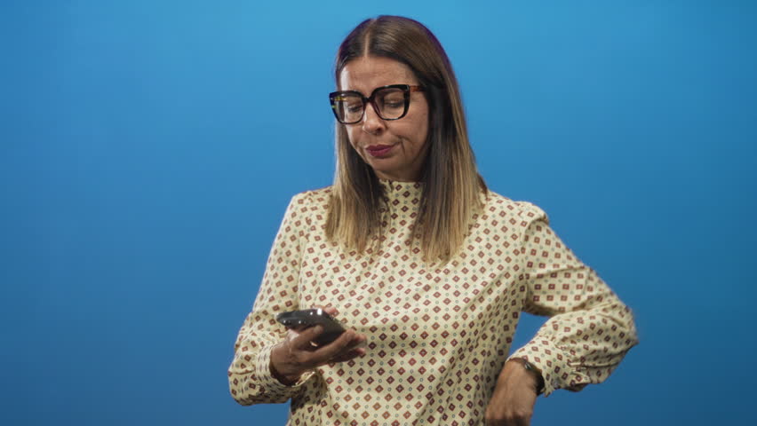 Woman holding smartphone with hand on hip checking the screen in studio, wearing patterned blouse and glasses; pensive concern.