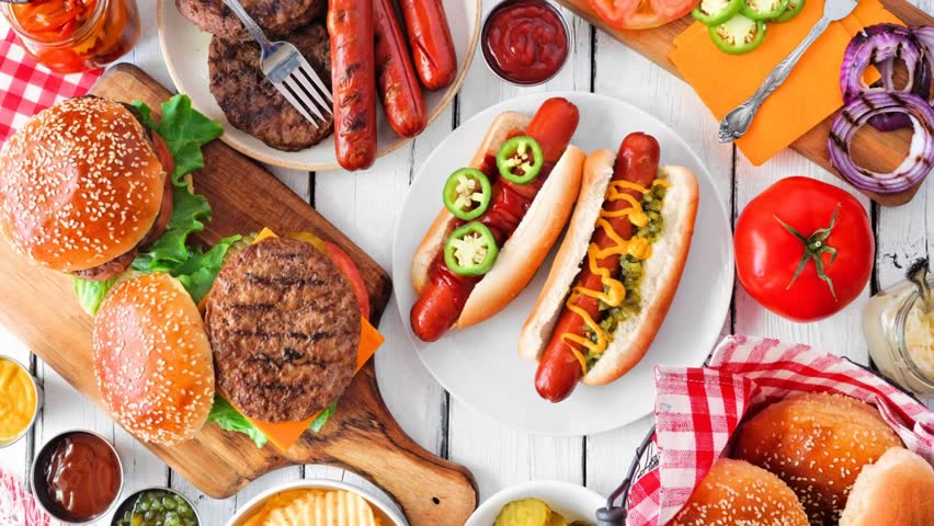 Summer BBQ food table scene with hot dog and hamburger buffet. Slow rotating zoom motion. Overhead view on a white wood background.