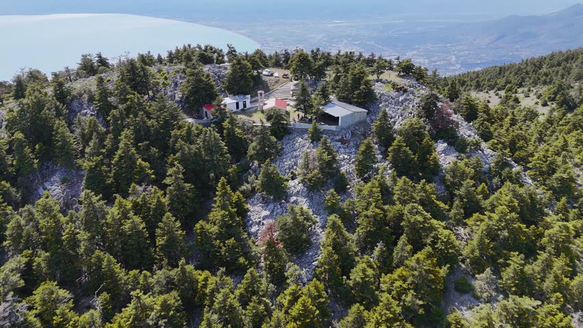 High-resolution aerial drone footage capturing a small church located on a mountain peak in the Peloponnese region of Greece, overlooking Kalamata and its coastline.

The composition combines natural and cultural elements: dense forested slopes, rocky terrain, and a hilltop structure that adds a strong focal point. In the background, the city stretches along the curved shoreline of the Mediterranean Sea, creating a layered and cinematic landscape.

The elevated perspective emphasizes isolation and tranquility, making the scene suitable for travel videos, documentaries, spiritual or cultural storytelling, and cinematic background visuals.

No identifiable people or brands visible.