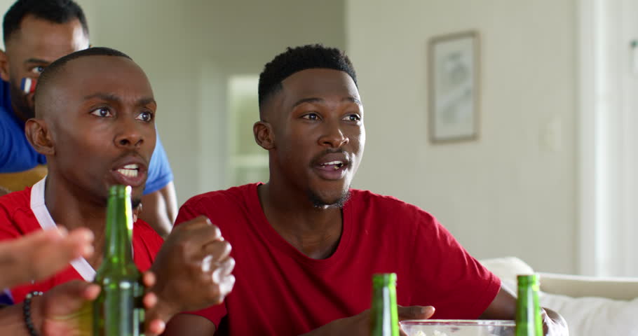 Three male friends in red and blue tees reacting to offscreen game, pointing, raising green bottle. Couch, popcorn, drinks, watching, reactions, camaraderie, casual