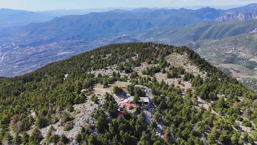 Aerial drone footage of a small chapel located on a forested mountain hill, surrounded by rocky terrain and trees, with a panoramic view over a coastal city and the gulf. The scene combines natural landscape with human presence, showing mountains, sea coastline, and urban area in the distance. Bright sunny weather, clear visibility, and wide cinematic perspective make this shot ideal for travel, nature, and tourism-related projects. Shot in Greece in high resolution.