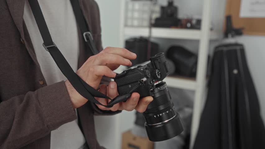 Man adjusting camera with hands and strap in studio, photographer checking lens, aperture and controls while standing near shelving and gear; quiet concentration.