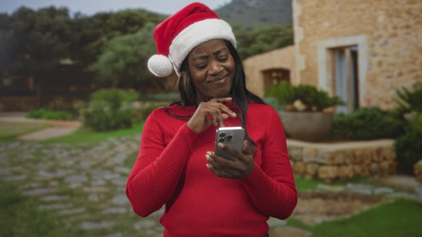 Woman wearing santa hat and red sweater, holding smartphone and tapping screen while smiling with hand on hip in front of stone building; holiday joy.