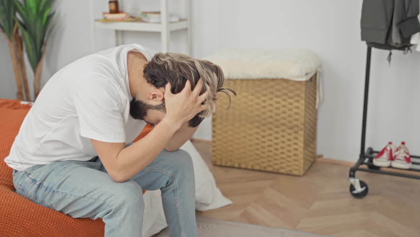 Man sitting on bed holding neck and head, clutching head with hands for headache in a building bedroom; anxiety stress fatigue.