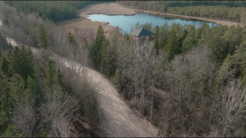 View of tower near lake surrounded by trees and dirt road in a forest area during daytime. Located in Latvia Lielie kangari nature park.