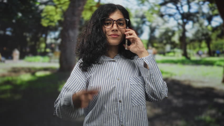 Woman holding smartphone to ear and shows two fingers to camera while wearing glasses and striped shirt in a forest park outdoors; playful confidence.