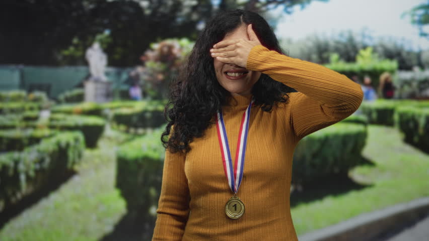 Woman young brunette wearing a gold medal on a ribbon, eyes closed and raising her hand on a street with trimmed hedges and a statue; quiet pride.