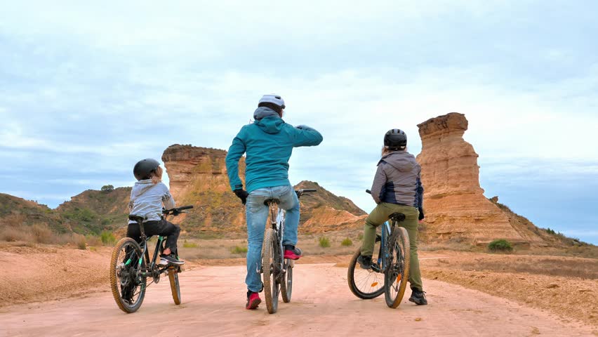 Family mountain biking adventure in Sierra de Armantes desert, Calatayud, Spain. Three cyclists on a gravel road looking at red rock formations and geological reliefs under cloudy sky.