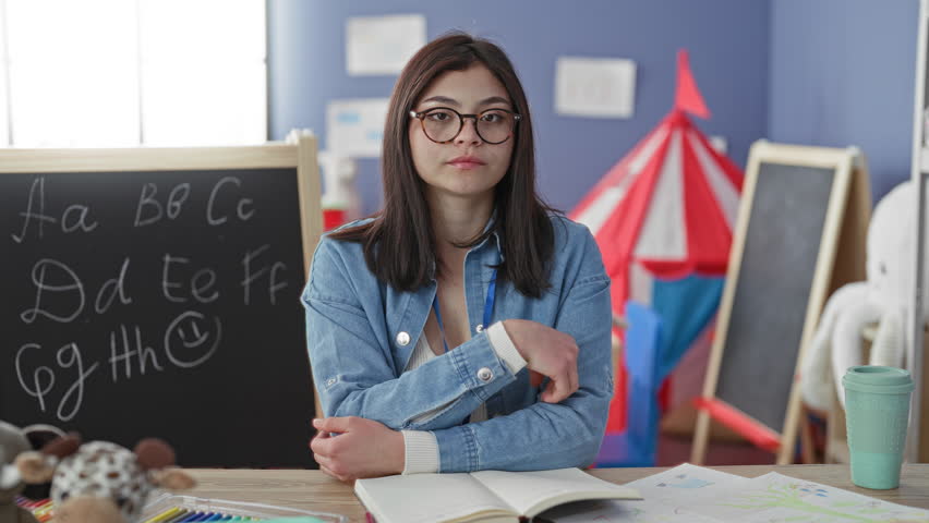 Woman teacher with open book at chalkboard, hand to mouth yawning over notebook and toys on table in classroom building; quiet fatigue.