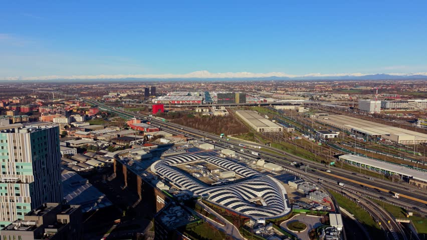 Expansive aerial view of milan, showcasing modern architecture and a bustling highway. The majestic snowy alps are visible on the horizon. Milan, Italy