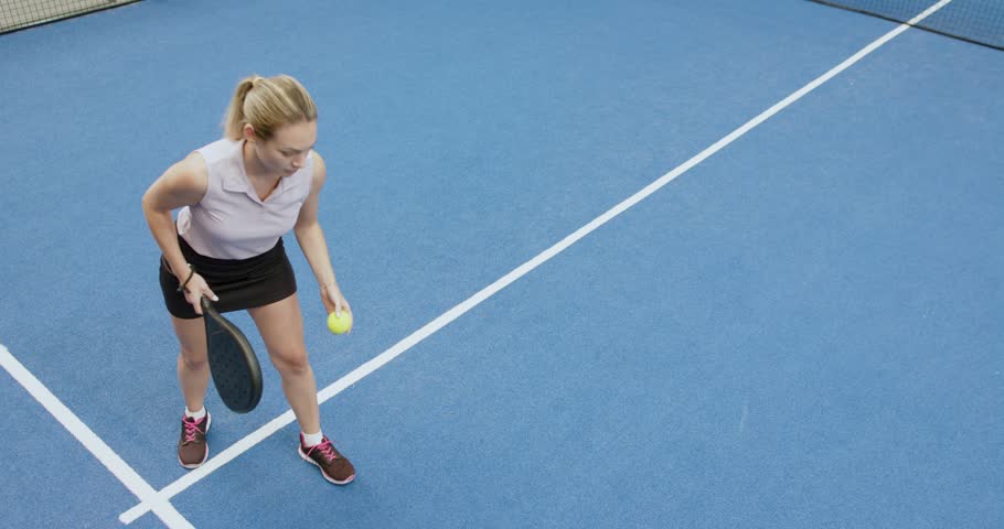 Female athlete bouncing ball to serve, tossing up, swinging paddle on blue court tracking flight. Sports, active, energetic, fitness, competition, training, agility